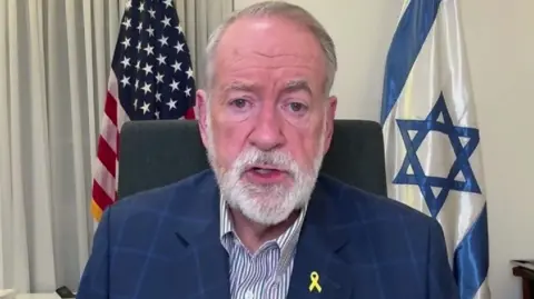 Mike Huckerbee sat in a suit in front of US and Israeli flags