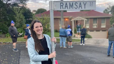 A woman smiles as she holds the pole of a street sign which says "Ramsay St", while seven people, some of them in blue beanies, stand in the street behind her. In the background is a suburban house