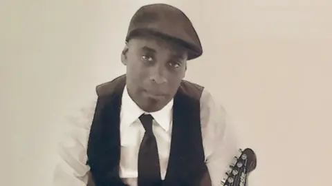 A sepia toned photo of Paul Reid sitting on a chair. He is wearing a flat cap, white shirt, black tie and black waistcoat with black trousers and shoes. He is looking into the camera and has a black and white guitar leaning on him.