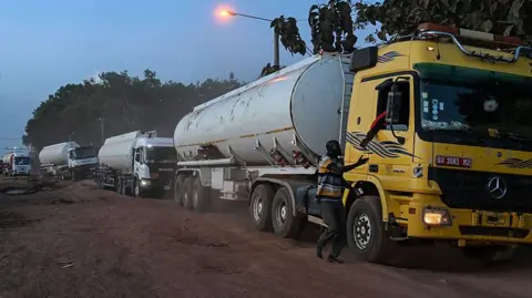 A line of large Malian fuel tankers stretches along a dusty road at dawn or dusk, their headlights and a streetlamp glowing through a hazy sky. A man walks beside the first yellow truck, holding on to its door as the convoy waits.