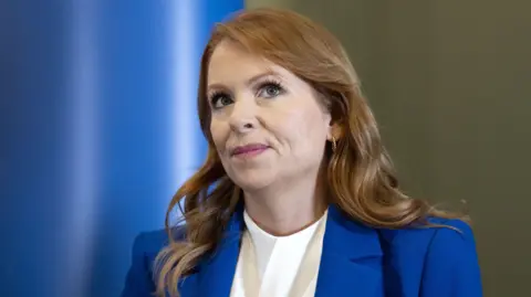 Ash Regan wearing a blue blazer over a white top. She has red hair falling over her shoulders. She is looking up, off camera.