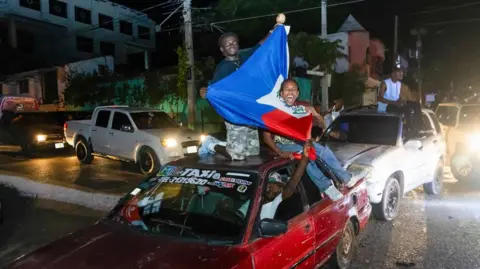 Two men on top of car holding Haiti flag