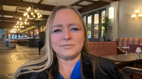 Shows a woman in blond hair with a blue blouse and black jacket standing in a hotel lobby