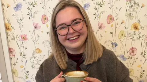 A blonde haired woman with tortoise shell glasses sitting smiling at the camera holding a mug of coffee. Behind her is floral wallpaper and she is wearing a brown knit cardigan.