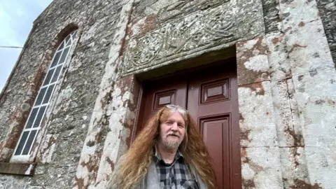 Archaeologist Andrew Nicholson stands in a doorway at the church, beneath an 8th Century stone lintel from Celtic times. He has long, red hair and glasses on his head. He is wearing a grey jacket and a black and grey checked shirt.