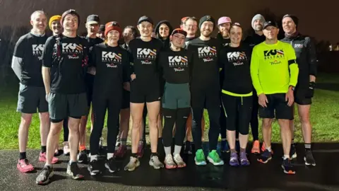 A group of runners standing together. It is raining and night time. Most of them are wearing black T-shirts which say "Run Belfast" on them in white lettering, one man to the front left of the group has the same logo but in black text on a neon yellow running top.