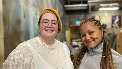 Jayde Adams wearing a white vest top over a white jumper stands next to Adryne Caulder-James at Bristol Old Vic. Adryne Caulder-James is wearing a light grey tracksuit top and has long dreadlocks