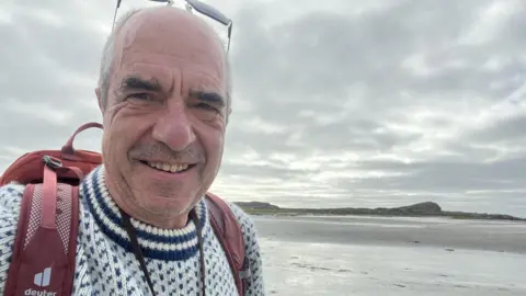 Steve Dickinson Steve Dickinson. He has short grey hair and is wearing a white and blue knitted jumper. He is standing on the Cumbrian coast. A hill can be seen in the distance.