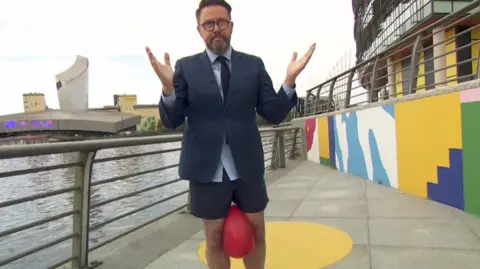 Jon Kay, in shorts has a red balloon between his knees, dressed from the waist up in a suit, shirt and tie