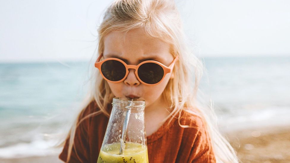 A young girl scowling, wearing sunglasses and sipping a glass of juice at the beach (Credit: Getty Images)